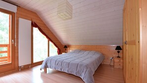 Una camera da letto con soffitto spiovente, pareti in legno, un letto con copripiumino grigio, due lampade da comodino e una finestra con tende rosse.