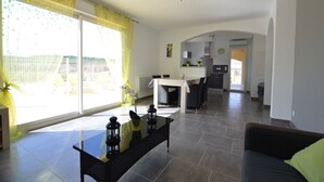A spacious living room with a large window, a dining area, and a kitchen in the background.