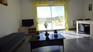 A living room with a sofa, a TV, a coffee table, and a view of the outside through a large window with yellow curtains.