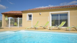 A pool area with a clear blue pool, a building with a tiled roof, and several green lounge chairs.