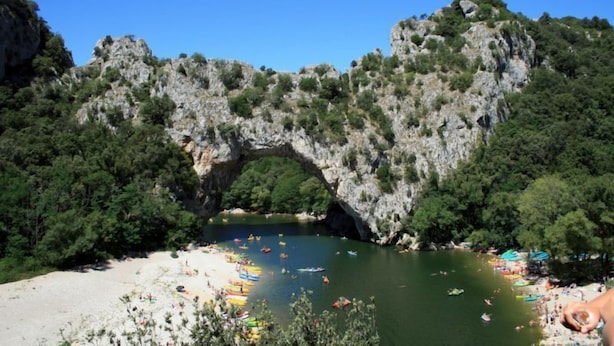 A natural rock arch over a river with boats and people on the shore.