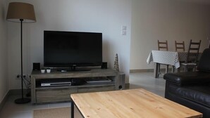 Una sala de estar con una televisión de pantalla plana sobre un soporte de madera, una lámpara, una mesa de comedor con sillas y un sofá de cuero negro.