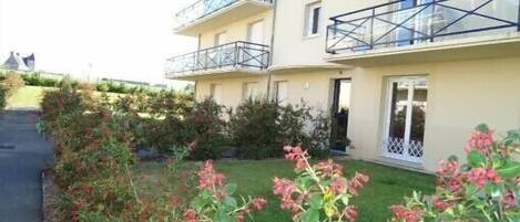 A multi-story residential building with balconies, a paved driveway, and a well-maintained garden with flowering plants.