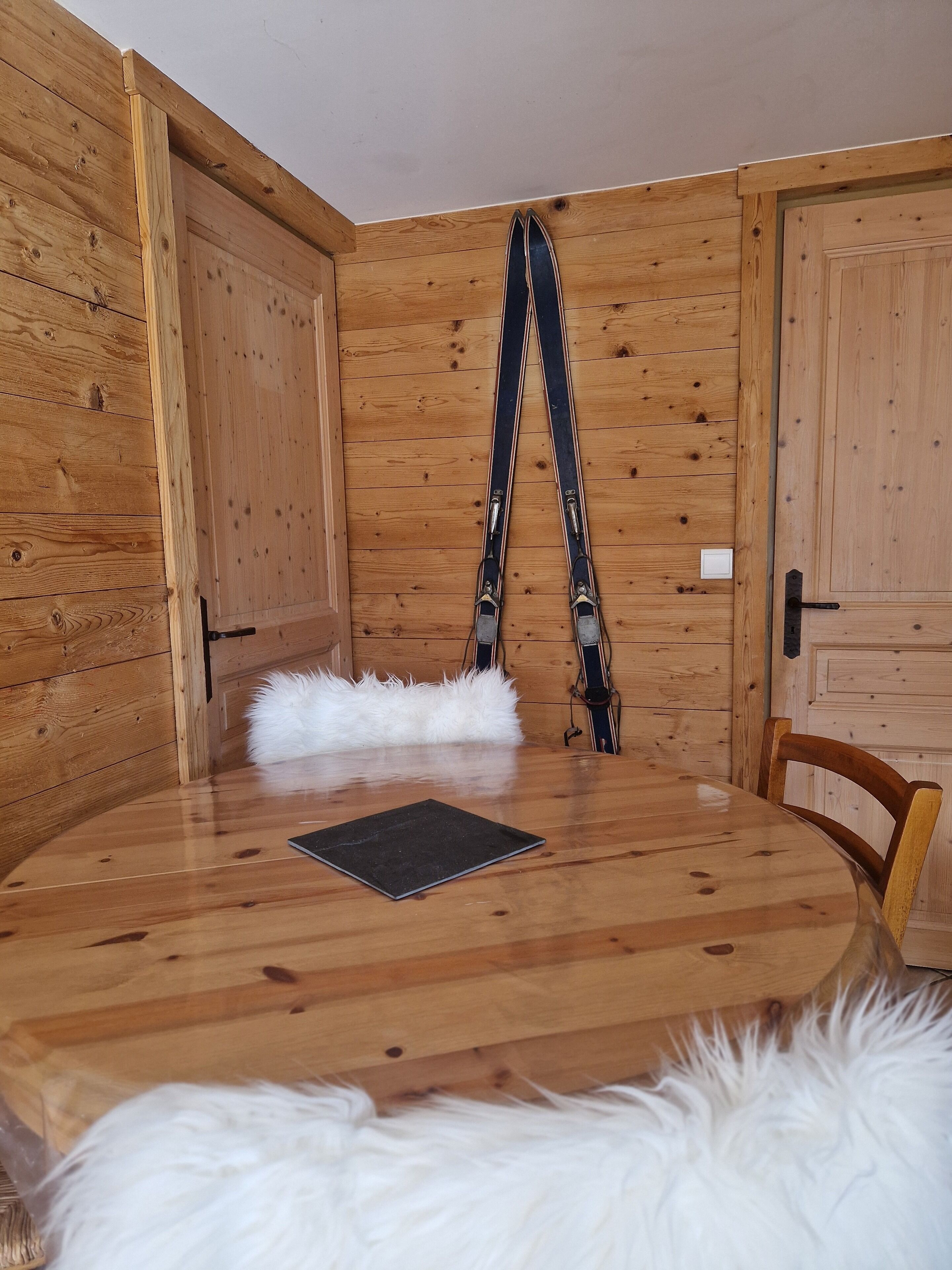 Une pièce en bois avec une table ronde, un tapis blanc et moelleux, et des paires de skis appuyés contre le mur.