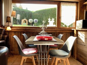 A dining area with a table set for four, surrounded by chairs, a lamp, and a view of a garden through a window. - GARDEN GROUND IN CHALET WITH WIFI CLASS 3 STARS (Chamonix-Mont-Blanc)