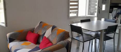 A living room with a sofa, a dining table with chairs, a clock on the wall, and a window with blinds.