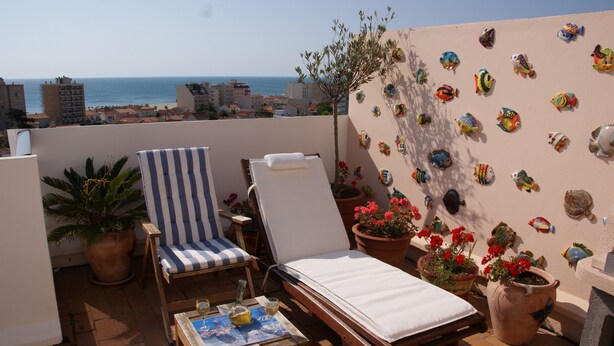 Une terrasse sur le toit avec deux chaises longues, une table avec une bouteille et un verre, et une décoration murale représentant des poissons.