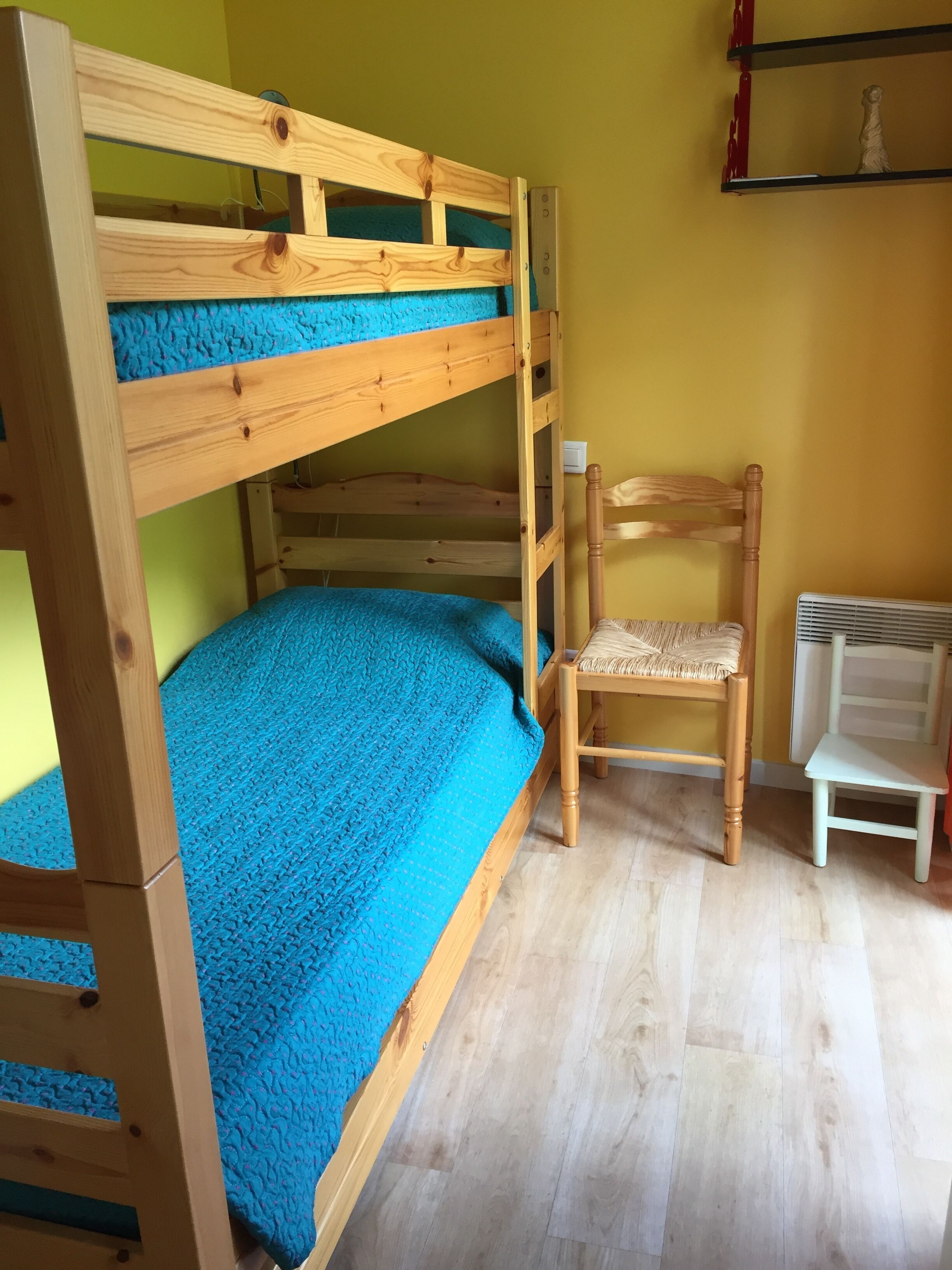 A bunk bed with a blue mattress, a wooden chair, and a white chair in a room with wooden flooring and a yellow wall.