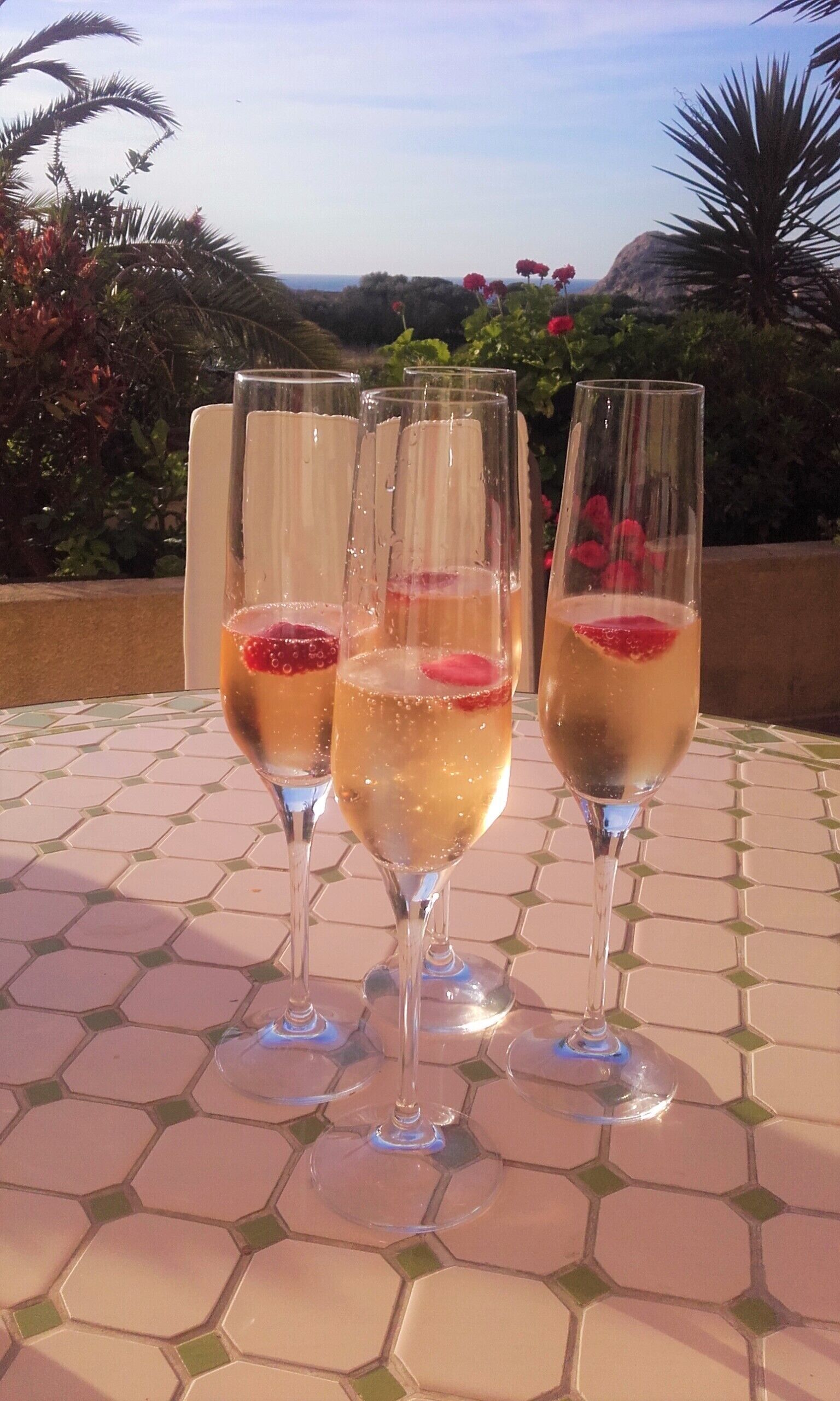 Tres copas de champán con adornos de fruta sobre una mesa de baldosas al aire libre.