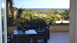 A patio with wicker furniture and a table, overlooking a lush landscape with trees and a view of the sea.