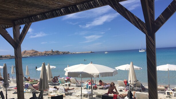 A beachside café with outdoor seating, umbrellas, and a view of the sea and boats.