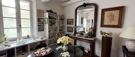 Un comedor con una mesa redonda, sillas, un jarrón con flores, una estantería con adornos y un espejo grande.