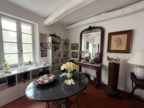 Een eetruimte met een ronde tafel, stoelen, een vaas met bloemen, een plank met decoratieve objecten en een grote spiegel.