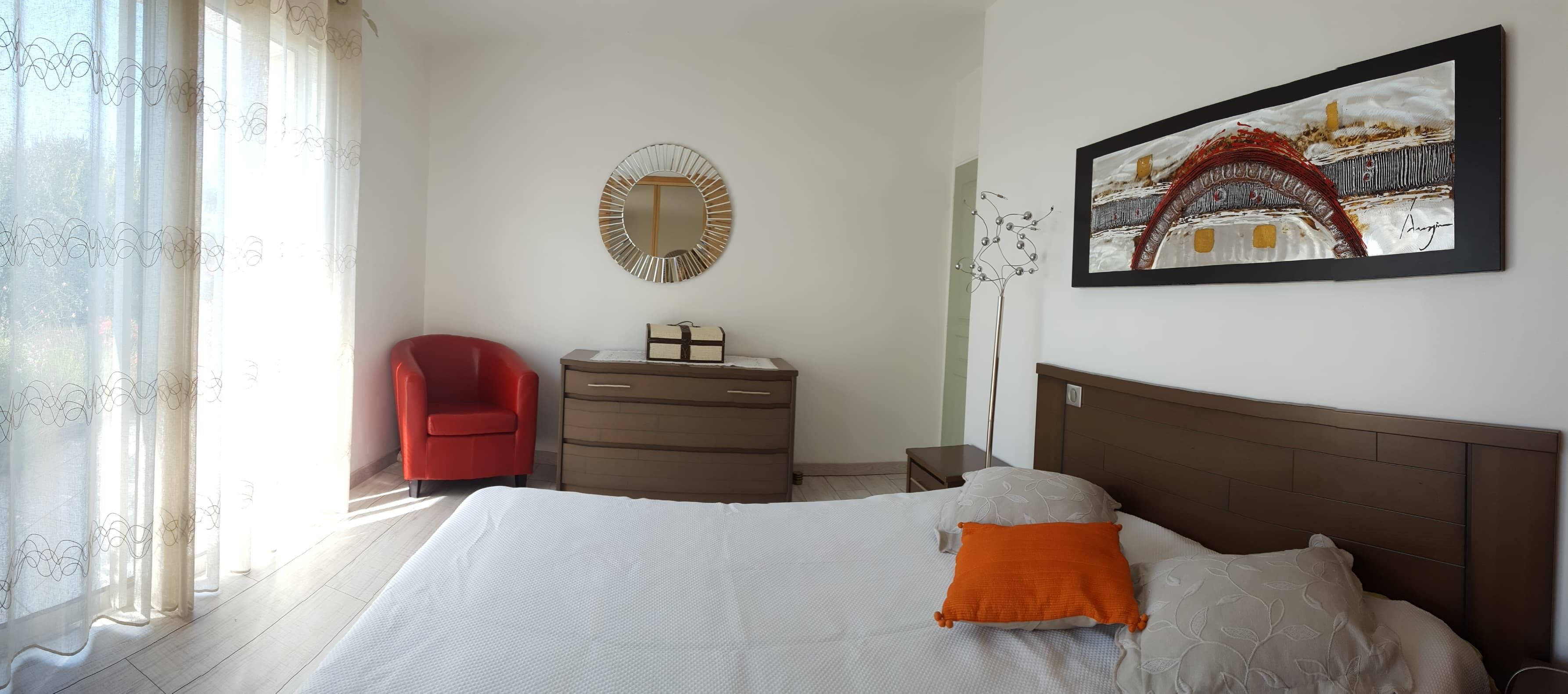 A bedroom with a bed, a red chair, a dresser, a mirror, and a framed picture on the wall.