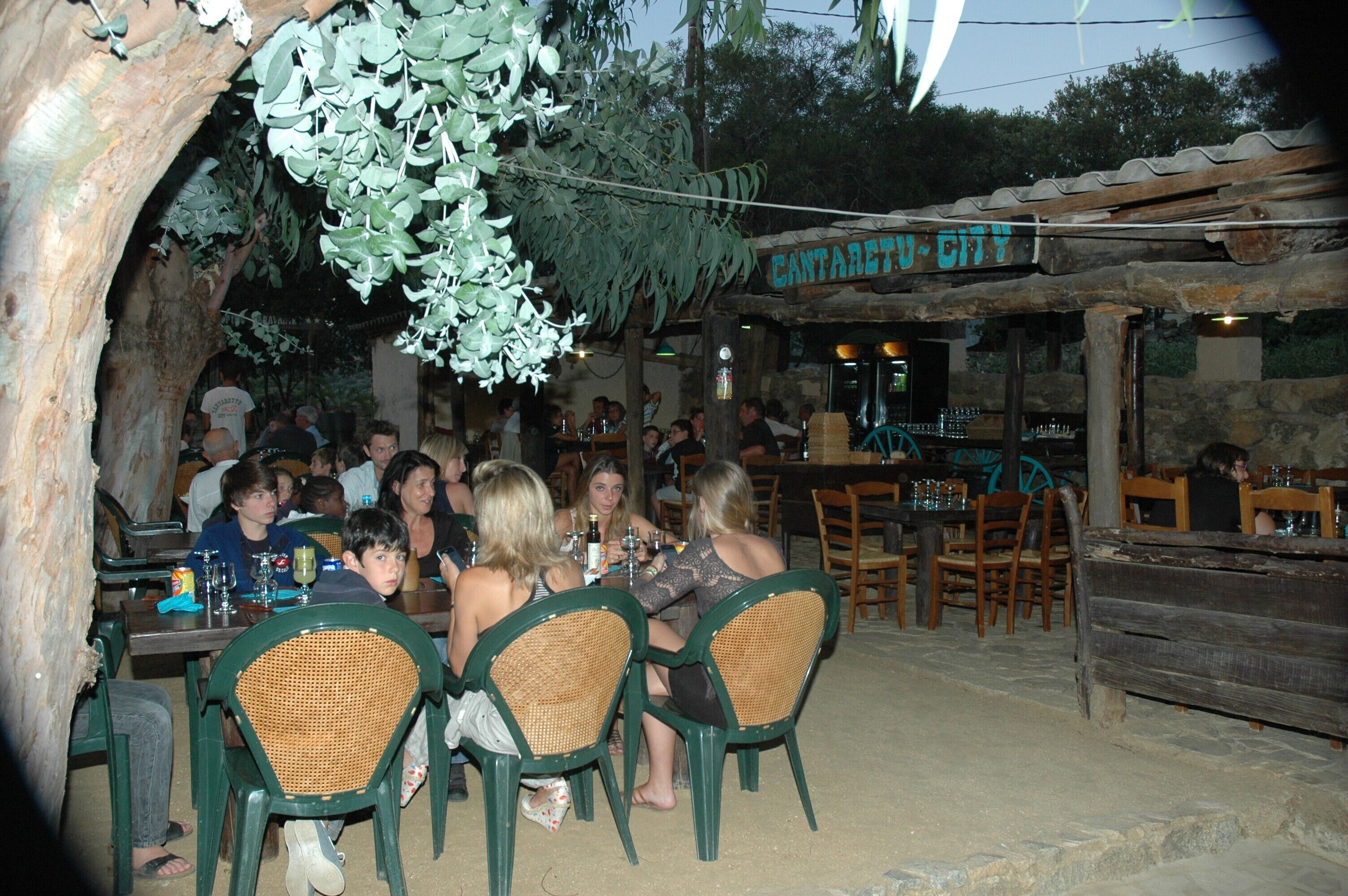 A rustic outdoor dining area with wooden tables and chairs, surrounded by greenery and a stone wall.
