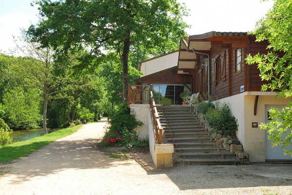 A wooden house with a stone staircase leading to a door, surrounded by greenery and a gravel path.