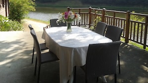 Un espacio de comedor al aire libre con una mesa preparada para dos personas, rodeado de sillas de mimbre y vistas a un cuerpo de agua y un área boscosa.