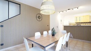 A modern dining area with a wooden table, white chairs, a hanging light fixture, and a kitchen in the background. - Gîte de la truffière "Les Chênes  (Montagnac-Montpezat)