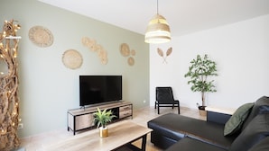 A modern living room with a flat-screen TV, a wooden coffee table, a black leather sofa, a black chair, and a hanging light fixture. - Gîte de la truffière "Les Chênes  (Montagnac-Montpezat)