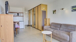 A modern living room with a sofa and a small table, a kitchen area with cabinets and a coffee machine, and a sliding door leading to another room. - HOSSEGOR Océan, studio for 3 people (HOSSEGOR)