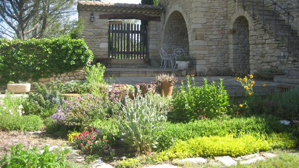 A stone building with a garden in front, featuring a wooden gate, stone arches, and various plants.