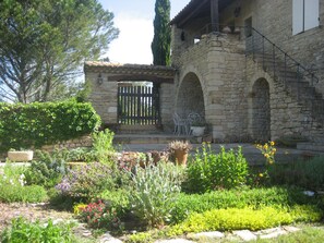 Un bâtiment en pierre avec un jardin à l’avant, agrémenté d’un portail en bois, d’arcs en pierre et de diverses plantes.