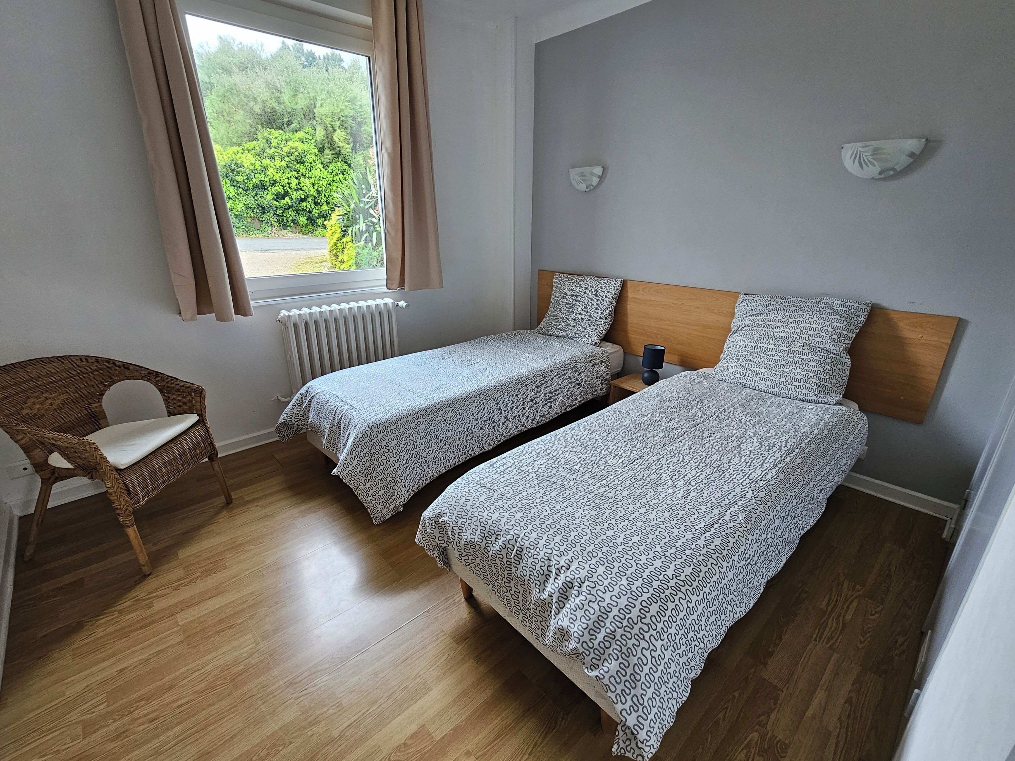 A room with two single beds, a wooden chair, a bedside table with a lamp, a window with curtains, and a radiator.
