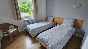 A room with two single beds, a wooden chair, a bedside table with a lamp, a window with curtains, and a radiator.
