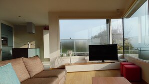 Una sala de estar moderna con un sofá, un televisor, una mesa de madera y una cocina al fondo.