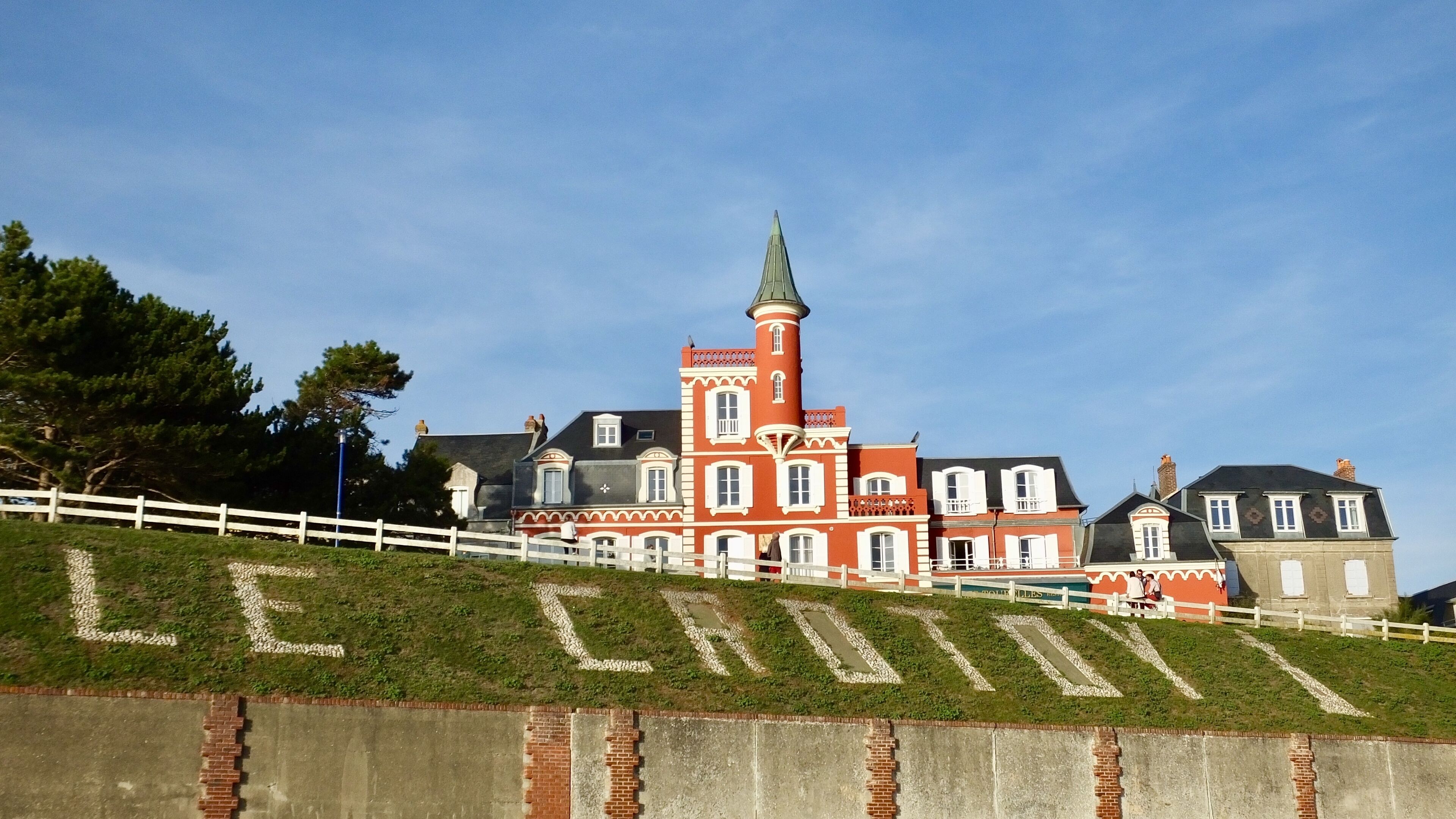 Un grand bâtiment historique, doté d’une tour imposante et de nombreuses fenêtres, situé sur une pente herbeuse, avec une clôture blanche en premier plan.