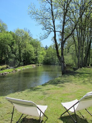 Une vue sur la rivière, avec deux chaises de jardin placées face à l’eau, le tout entouré d’une végétation luxuriante et d’arbres.