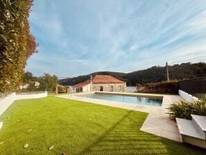 A swimming pool with a stone house in the background, surrounded by a well-maintained lawn and a hedge.