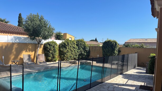 Une piscine dotée d’une plage en pierre, entourée d’une clôture et jouxtant un bâtiment dont le toit est recouvert de tuiles.