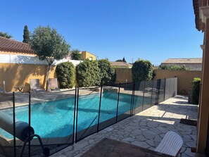 Una piscina con plataforma de piedra, rodeada por una valla y adyacente a un edificio con techo de tejas.