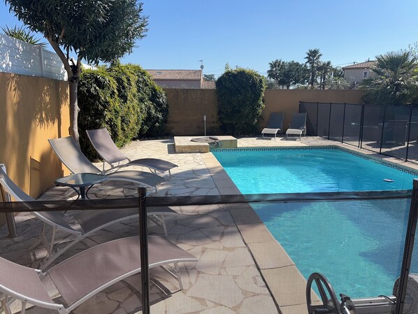 Un área de piscina con sillas de descanso y una pequeña mesa, rodeada por una valla y follaje.