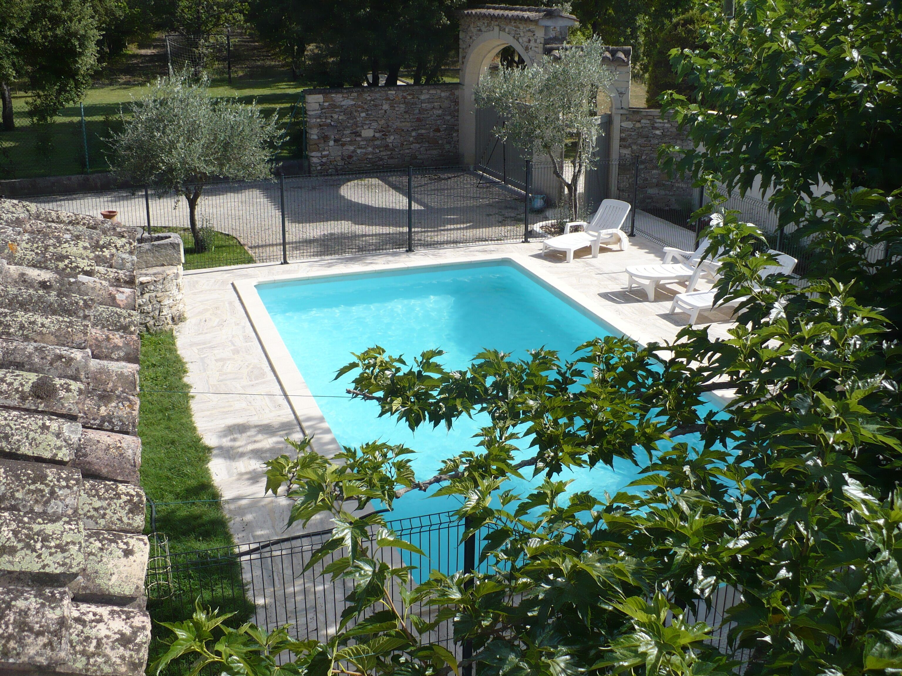 A rectangular swimming pool with a stone wall and a metal fence. There are white lounge chairs and a small stone archway.