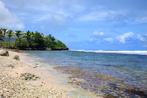 A beach with clear water, pebbles, and a small island with trees and a building.