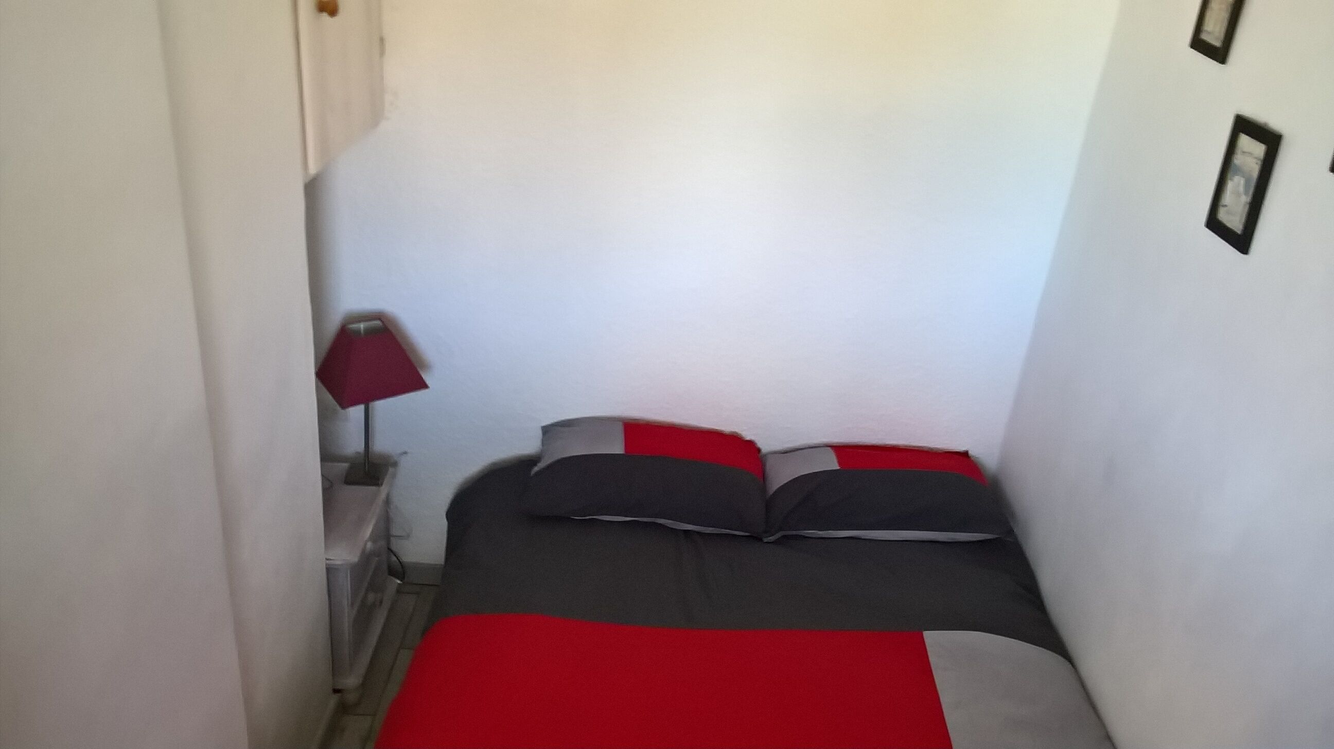 Ein kleines, schmales Zimmer mit einem Bett, einem Nachttisch und einer roten Lampe.