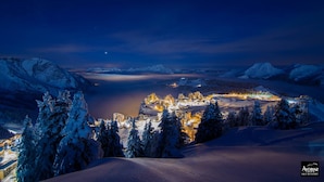 Paisaje montañoso nevado de noche con edificios iluminados y una vista despejada de un lago.