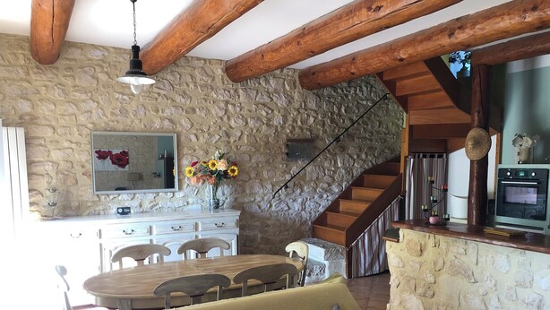 Una cocina rústica con paredes de piedra, vigas de madera, una zona de comedor con mesa y sillas, y un sofá con cojines.