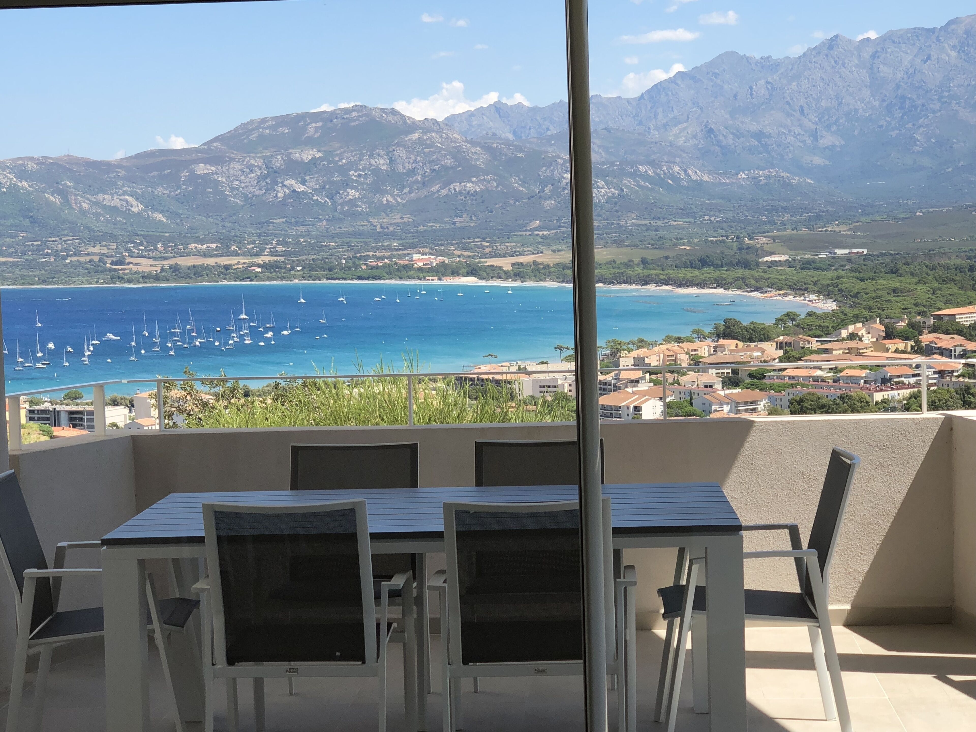 A balcony with a table and chairs, overlooking a coastal area with boats and mountains.