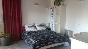 Slaapkamer met een bed, twee kussens, een nachtkastje, een lamp, een radiator, een kledingkast, een potplant en een rood gordijn.