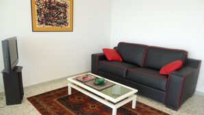 Ein Wohnzimmer mit einem schwarzen Sofa, roten Kissen, einem Glastisch, einem gerahmten abstrakten Gemälde und einem Fernseher auf einem Schrank.
