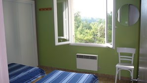 Una habitación con dos camas, una ventana con vista a árboles, una silla blanca y un espejo redondo en la pared.