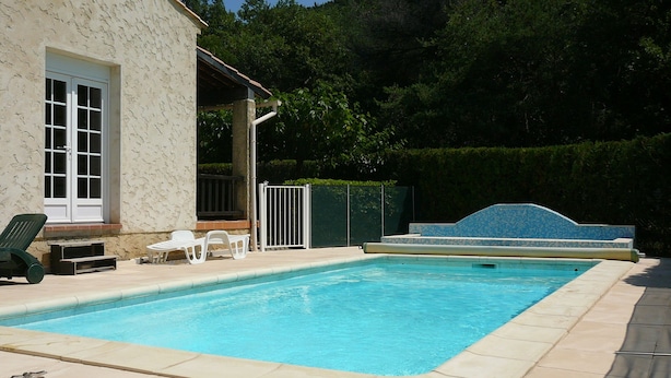 Una piscina rectangular con solárium empedrado, un porche cubierto con mobiliario exterior y un muro de piedra.