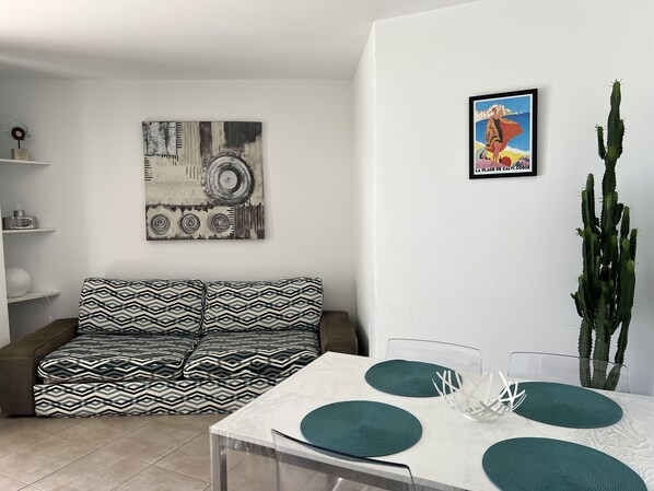 A modern living room with a patterned sofa, a dining table with green placemats, a cactus, and artwork on the walls. - CALVI, 1-bedroom apartment in a residence with ocean views, Wi-Fi, 5 minutes from the beach and downtown, air-conditioned, sleeps 4 (Calvi)