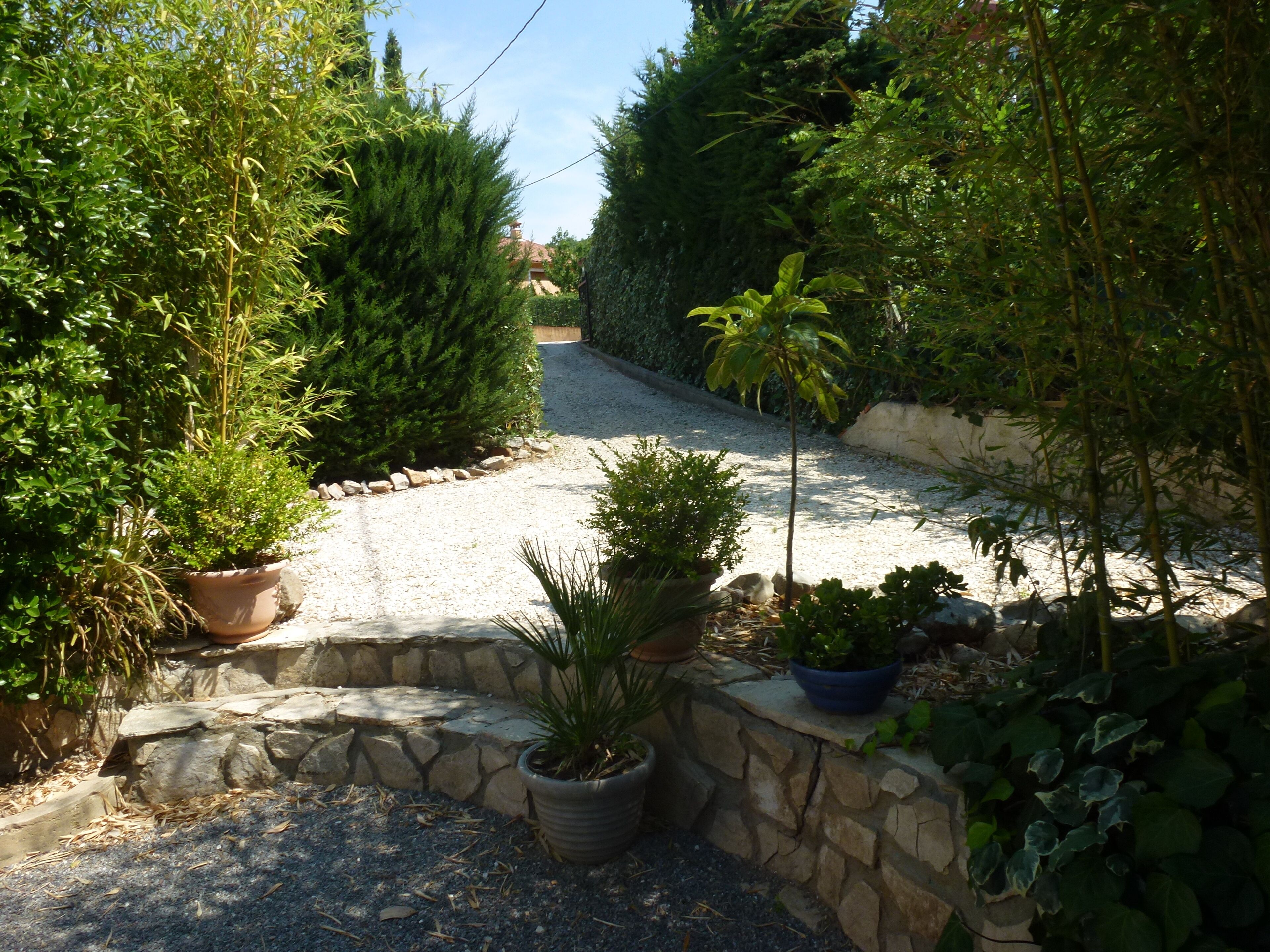 Un camino de grava que lleva a una casa con un muro de piedra y plantas en macetas.