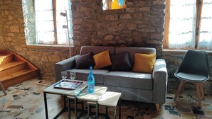 Une pièce chaleureuse avec un mur en pierre, un canapé gris agrémenté de coussins orange, une petite table avec deux verres et une bouteille, et un fauteuil.