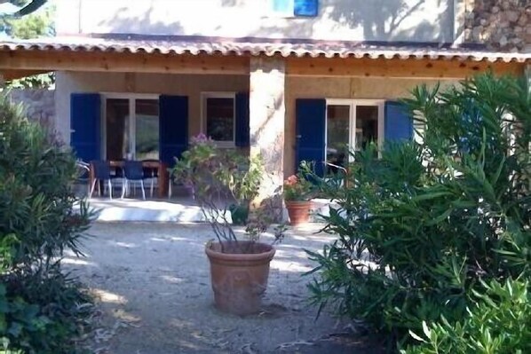Un edificio rústico con tejado de tejas y contraventanas azules. Hay plantas en macetas en primer plano y una silla afuera.