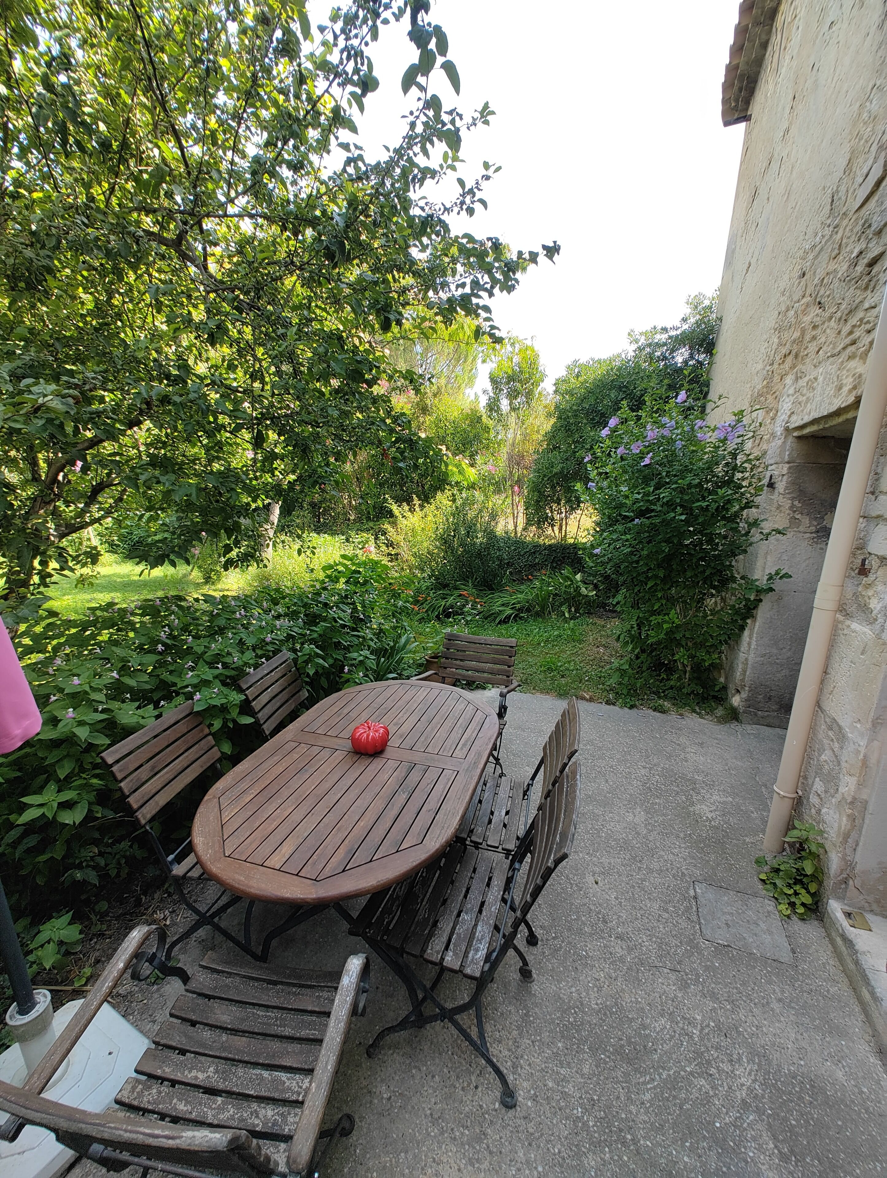 An outdoor patio with a round wooden table and benches, surrounded by lush greenery and a stone wall.